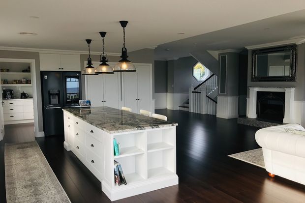 Open-plan kitchen and living area with a large white island featuring a dark patterned stone benchtop, white shaker-style cabinetry, black appliances, pendant lighting, and dark timber flooring. The adjoining living room includes a fireplace with a wall-mounted television, while a staircase and hallway are visible in the background.