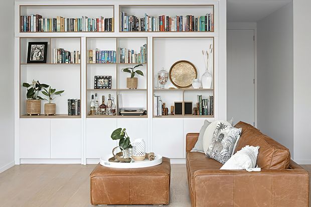 Bright, carefully styled living room with a built-in white shelving and cabinetry wall displaying books, plants, framed artwork, glassware, and decorative objects. A cognac brown leather sofa and matching ottoman sit on light timber flooring, with neutral cushions and a round tray creating a warm, refined reading and lounging area.