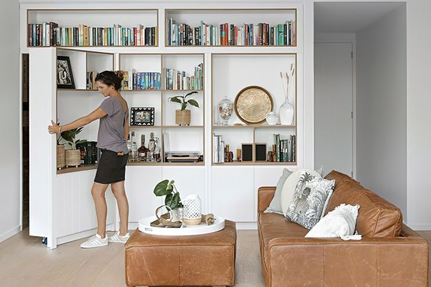 Bright, refined living room featuring a full-height built-in white shelving and cabinetry wall filled with books, decor, plants, and a small bar display. A warm brown leather sofa and matching ottoman sit on pale flooring, with a woman opening a concealed or integrated storage section. The room has a clean, tailored, contemporary feel with subtle classic detailing in the shelving trim.