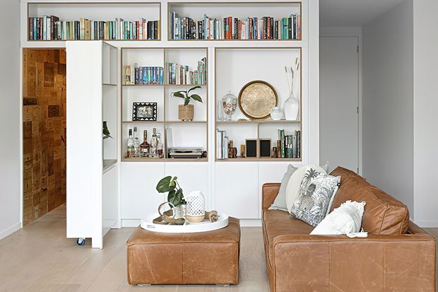 Bright, refined living room with a large cognac leather sofa and matching ottoman, white built-in shelving filled with books and decorative objects, and a white oval coffee table. A concealed or pivoting shelving panel opens toward a timber-lined passage, while a plain white door and light flooring complete the space.