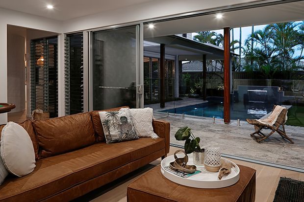 A warm contemporary living room with a large brown leather sofa and matching ottoman coffee table, opening through extensive sliding glass doors to a covered outdoor entertaining area and swimming pool. Timber posts, stone paving, indoor plants, and louvred glazing create a strong indoor–outdoor connection.