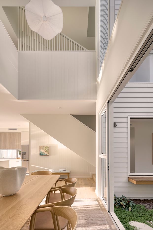 Bright, refined interior view centred on a timber dining table and curved wooden dining chairs beside large open glazing. The space features light vertical wall panelling, a staircase with white balustrades, a double-height void, a large sculptural pendant light, and a view toward a small planted courtyard and weatherboard-clad exterior.