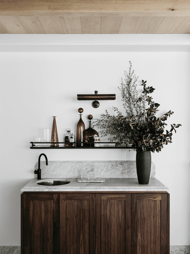 A refined compact kitchenette or bar area with dark timber cabinetry, a pale marble benchtop and short backsplash, black gooseneck tap, undermount sink, open metal display shelf, and an aged bronze wall light. Decorative glassware, bottles, and a tall vase of foliage sit on the shelf and counter. A light timber ceiling adds warmth to the otherwise crisp white setting.