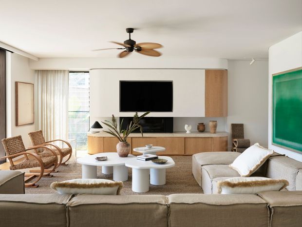 A bright, warm living room with a large upholstered sectional sofa, woven wood lounge chairs, layered round white coffee tables, and a built-in timber and white media wall. Floor-to-ceiling curtains and glazing bring in daylight, while a timber-bladed ceiling fan and natural decorative objects add organic texture. A large green artwork provides the main accent colour.