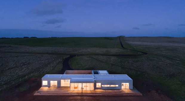 Aerial dusk view of a low, elongated modern single-storey house set in a broad, sparsely vegetated rural landscape. The building has a simple rectangular form, a shallow roof, extensive illuminated glazing, and a timber-toned deck or terrace extending along the front.