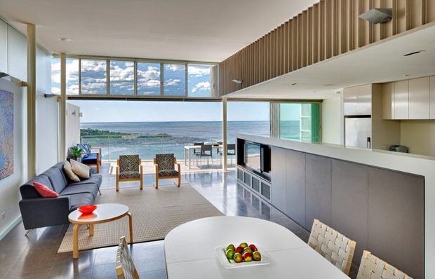 Bright open-plan coastal living and dining space with a contemporary kitchen, extensive glazing, and direct ocean views. The room features a dark sectional sofa, pale wood and white furnishings, a large white dining table, a streamlined grey kitchen island, polished flooring, and a vertical timber slat feature wall extending toward the ceiling.