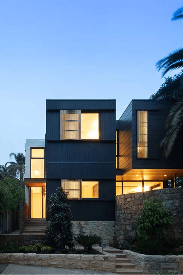 A contemporary multi-level residence photographed at dusk, composed of dark vertical-clad box forms with warm illuminated windows and interiors. Stone retaining walls, landscaped planting, stairs, and palm trees frame the front entry, while a projecting upper volume creates a sheltered outdoor area.