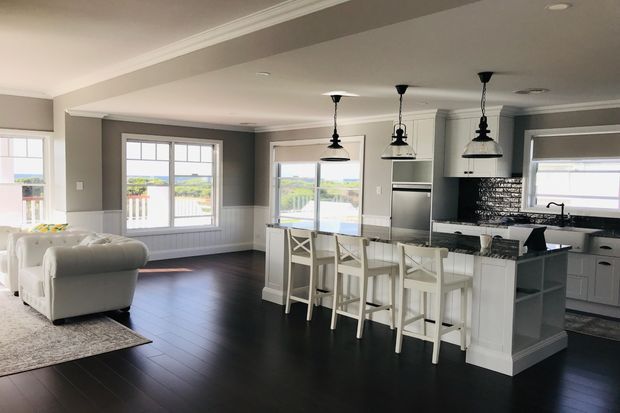 Open-plan kitchen and living area with dark timber flooring, grey walls, white cabinetry and trim, a large central island with seating for four, black pendant lights, and expansive windows providing bright outdoor views. The living area includes white upholstered sofas and a light rug.