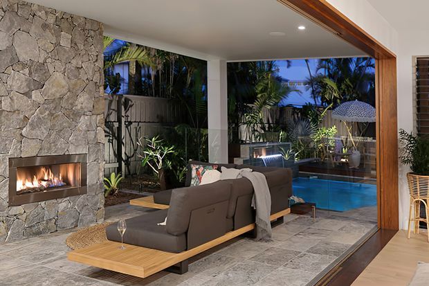 A sophisticated open-plan indoor-outdoor living area at dusk, centred on a large charcoal sectional sofa facing a built-in linear fireplace clad in irregular natural stone. The room has large open glass doors framed in warm timber, grey stone-look flooring, tropical planting, and a visible blue swimming pool and outdoor seating area beyond. A timber-framed opening connects the living room to the outdoor space.