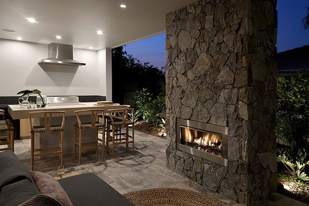 An open-plan indoor-outdoor entertaining space at dusk, combining a compact contemporary kitchen with a large island and timber bar stools, a partially visible living area, and a dramatic floor-to-ceiling rough stone fireplace wall with a built-in gas fire. Recessed ceiling lighting illuminates the kitchen and textured stone flooring, while garden planting and the dark exterior are visible through the open wall.