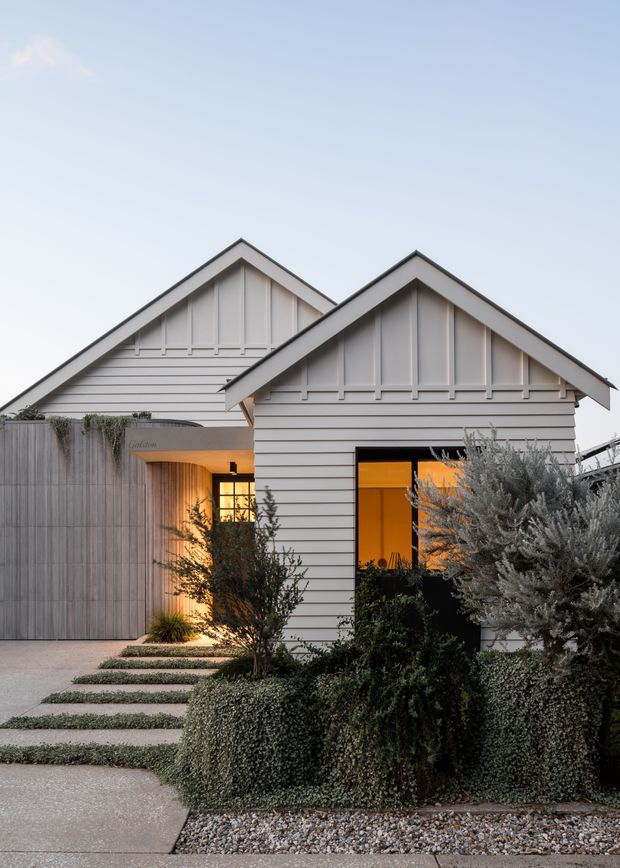 Front exterior view of a compact residential property composed of two overlapping gabled volumes. The facade is clad in pale horizontal weatherboards with contrasting vertical gable detailing and dark trim. A warm-lit recessed entry sits beside a tall dark-framed window, while layered drought-tolerant planting, clipped groundcover, gravel, concrete paving, and stepped garden paths define the landscaped approach.