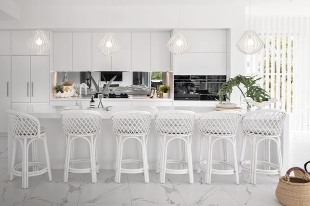 Bright white contemporary kitchen with a long island and five woven-back bar stools. The space features white paneled cabinetry, mirrored splashback, integrated appliances, pendant lighting, marble-look flooring, and vertical blinds with greenery visible beyond.