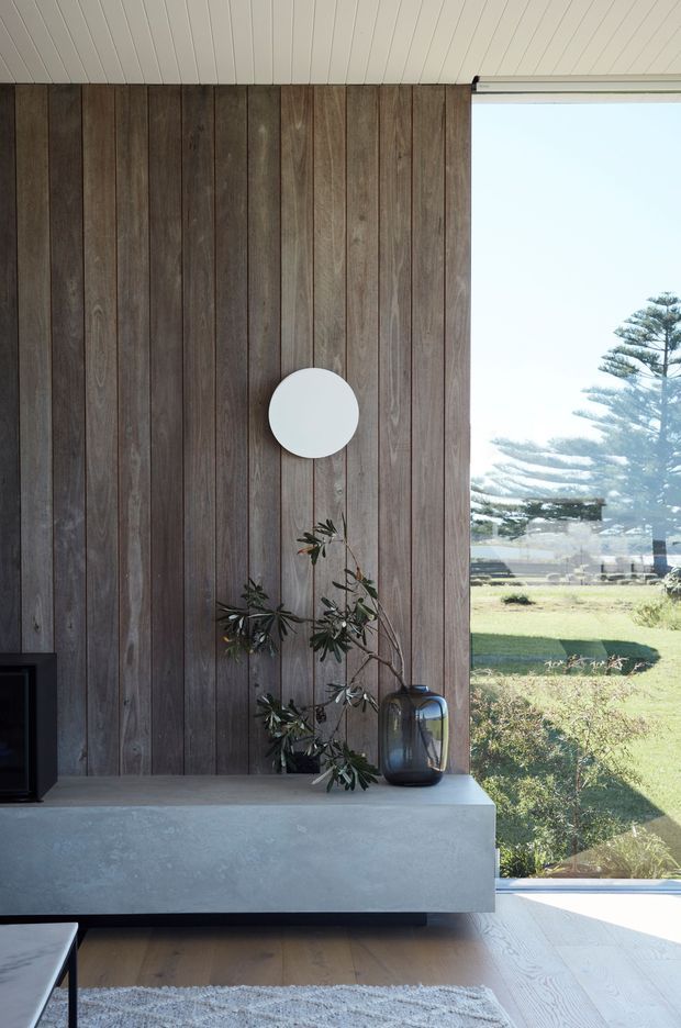 A calm, contemporary living room interior featuring a dark vertical timber feature wall, a minimalist concrete or rendered built-in media console, pale timber flooring, and a white linear-panel ceiling. A large window provides views toward a grassy coastal or rural landscape. Minimal decor includes a round wall light, a dark glass vase, and a small indoor plant.