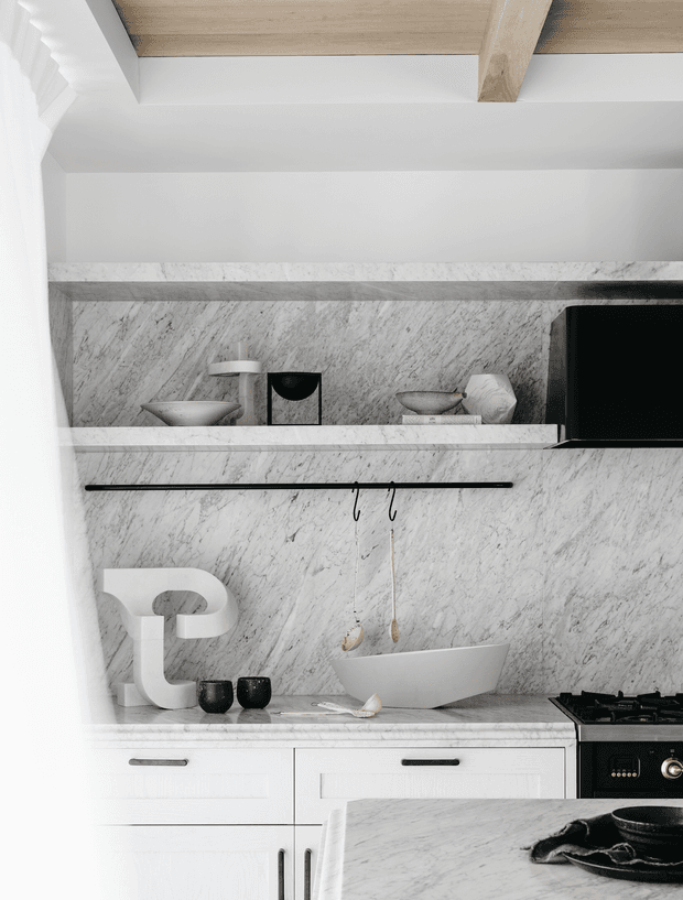 A refined white and grey kitchen with extensive veined stone wall cladding and benchtops, white lower cabinetry, open display shelving, and black hardware and appliances. A timber-lined ceiling with a prominent exposed timber beam adds warmth and architectural character. The styling is minimal and monochromatic with sculptural ceramics and restrained decor.