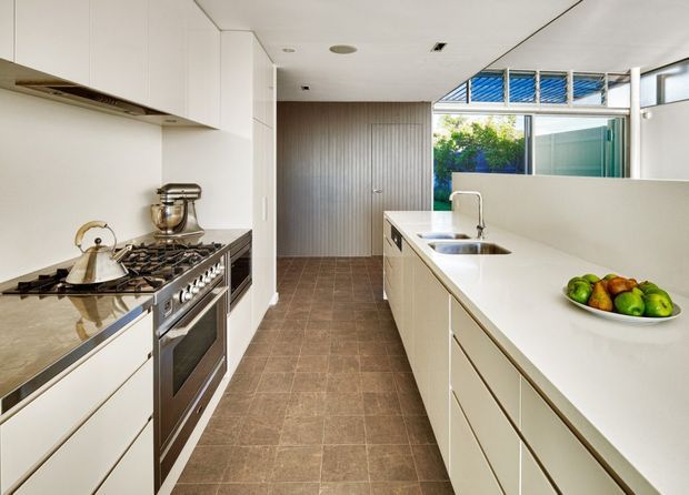 A bright, contemporary galley-style kitchen with extensive white flat-panel cabinetry, a long white island with integrated stainless-steel sink and mixer tap, and a stainless-steel range cooker beneath a built-in hood. The floor is finished in large brown-grey tiles, while a vertically ribbed grey feature wall and extensive windows and clerestory glazing bring in natural light. A fruit bowl and countertop appliances provide limited styling detail.