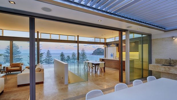 Open-plan contemporary living, dining and kitchen space with expansive floor-to-ceiling glazing overlooking a coastal landscape. The room features a timber kitchen island and cabinetry, stone flooring, a dining table with pendant lights, a lounge area, and a covered outdoor dining area in the foreground. A glazed skylight or operable roof structure and slim dark window frames create a bright indoor-outdoor connection.