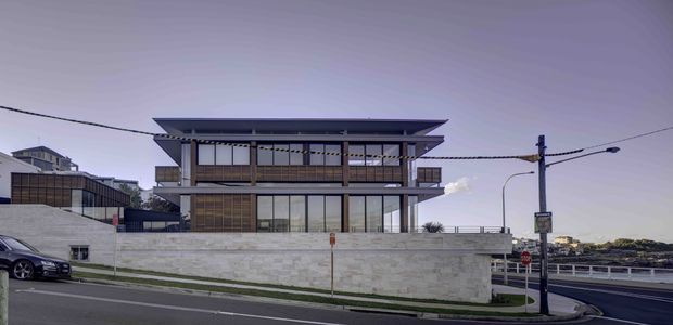 Street-level exterior view of a contemporary multi-storey building with broad cantilevered roof eaves, extensive glazing, dark vertical framing, and warm timber-toned screened sections. The building sits behind a pale masonry boundary wall on a corner site, with neighboring structures and roadway visible.