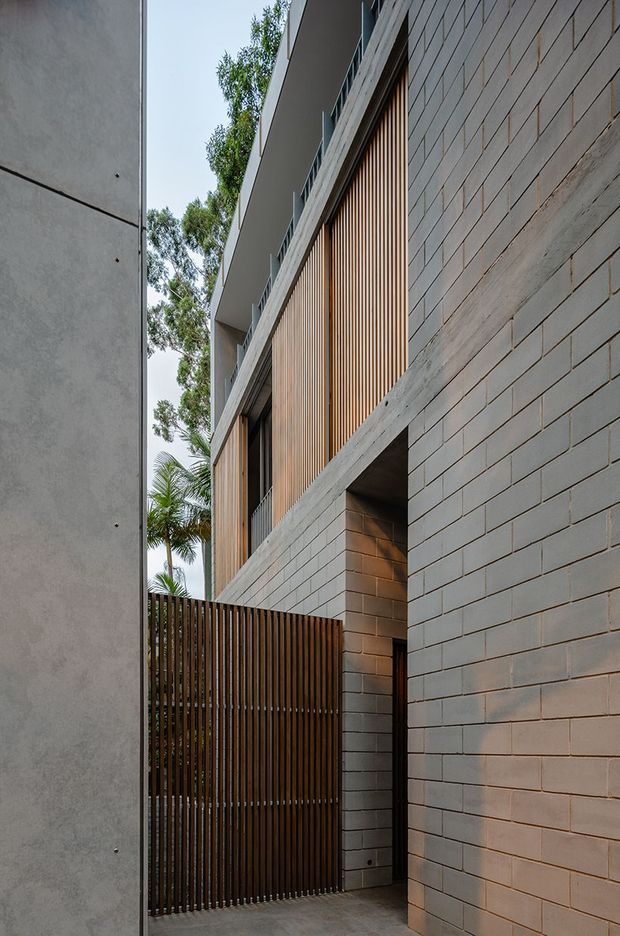 Narrow exterior passage between contemporary buildings, featuring pale grey masonry block walls, timber vertical screening and a matching timber slatted gate. The facade includes projecting concrete edges, dark recessed openings and a clean flat roofline, with subtropical planting visible in the gap. The composition is architectural and minimalist, emphasizing layered textures, privacy screening and a sheltered entry-like threshold.