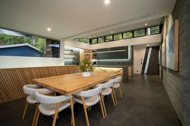 A contemporary open-plan dining and kitchen space with a large solid timber dining table surrounded by upholstered dining chairs. A built-in timber-slat bench runs along the left wall, while the kitchen features dark cabinetry, a long island or peninsula, integrated lighting, and a reflective splashback or feature wall. High clerestory windows bring in natural light, and a dark textured wall with large artwork occupies the right side. The room has polished concrete flooring and a warm, refined material palette.