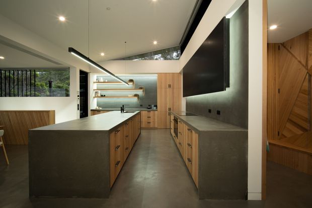 A contemporary open-plan kitchen with a large central island and parallel cabinetry, featuring dark concrete-look benchtops, warm timber joinery, integrated appliances, open timber shelves, and a large black feature wall with concealed television or display. The space has polished concrete flooring, recessed spotlights, linear pendant lighting, and generous glazing overlooking greenery. A timber-lined adjoining space is visible at right.