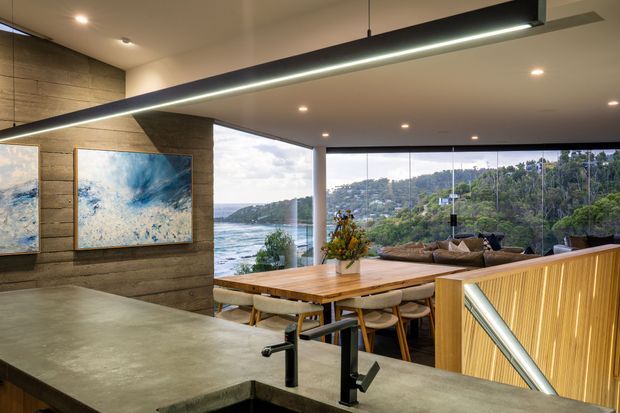 A contemporary open-plan kitchen and dining area with expansive floor-to-ceiling glazing overlooking a wooded coastal inlet. The foreground features a large grey stone island with matte black tapware, while a timber dining table, upholstered chairs and a lounge area sit beyond. A horizontal timber-clad feature wall displays artwork, and a timber slatted stair balustrade and linear pendant light add strong architectural detail.
