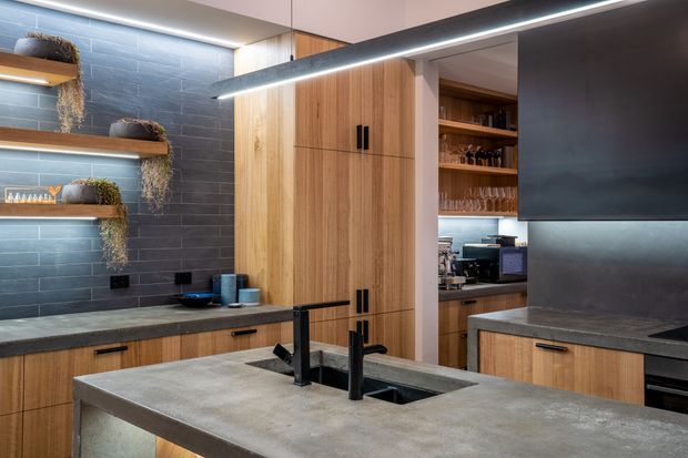Contemporary kitchen featuring a large concrete-look island with a black undermount sink and matte black tapware, timber-grain cabinetry, dark grey tiled splashback walls, open display shelving with plants and glassware, and integrated linear pendant lighting. A pantry or secondary storage area is visible through the opening at the rear.