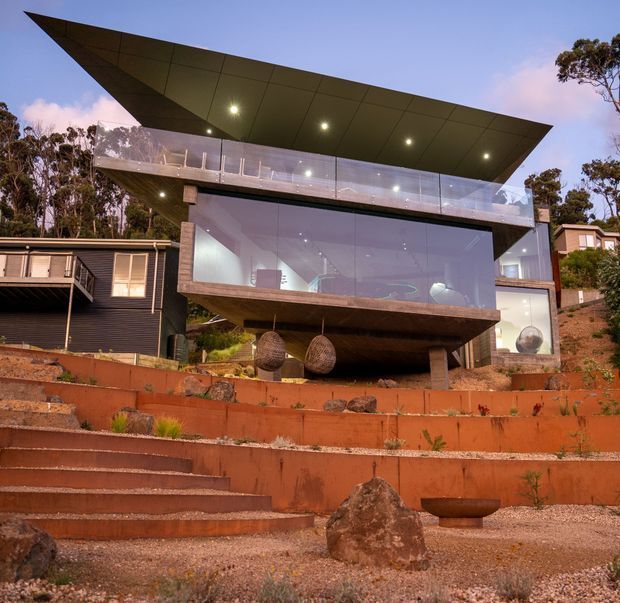 Exterior view of a striking contemporary multi-level house set into a landscaped, terraced site. The building features a dramatic cantilevered wedge-shaped roof, extensive floor-to-ceiling glazing, a projecting upper balcony with transparent balustrades, dark cladding, and exposed stone or concrete structural elements. Terracotta retaining walls, steps, gravel, boulders, and native planting form the foreground landscape.