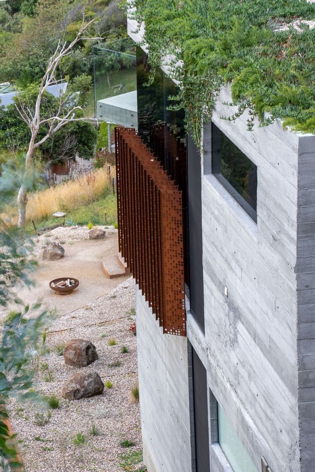 Exterior architectural view of a contemporary hillside dwelling, showing a strongly textured light-grey concrete wall, narrow dark-framed windows, and a projecting rusted steel privacy screen or sunshade. The building is integrated into a landscaped rocky garden with gravel, boulders, native vegetation, and a small fire bowl. Reflective glazing and dense trees frame the facade.