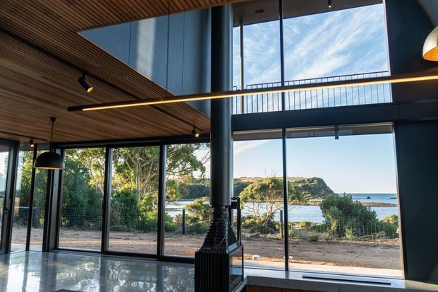 A dramatic, open-plan interior with floor-to-ceiling glazing overlooking a coastal bay. The space features a double-height void, dark structural columns, warm timber slatted ceiling, polished concrete flooring, black-framed windows, and linear pendant lighting. Dense vegetation and a rocky shoreline are visible outside.