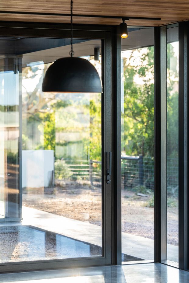 A contemporary interior corner with extensive floor-to-ceiling glazing and dark metal-framed glass doors opening toward a leafy outdoor area. The space has a polished concrete floor, a warm wood-panelled ceiling, and a large black dome pendant light, creating a modern indoor-outdoor connection.