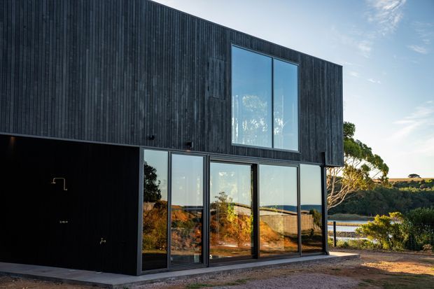 Contemporary two-storey house exterior clad in dark vertical timber boards, with expansive black-framed glazing on both levels. The building overlooks a landscaped rural or coastal setting with water visible beyond, and includes a recessed dark ground-level opening.