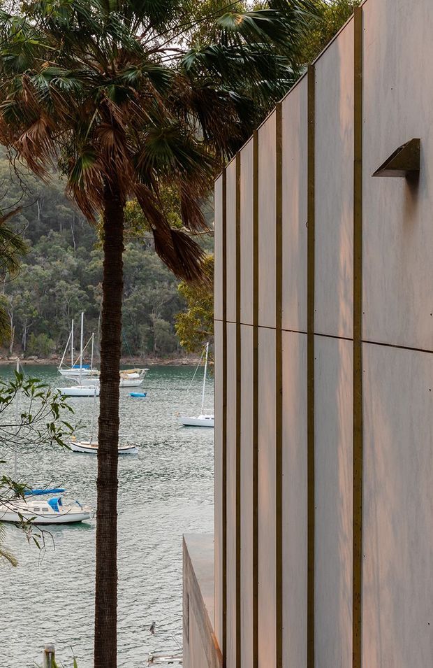 Exterior architectural detail beside a calm waterfront, featuring a tall pale rendered wall with evenly spaced vertical timber battens and a small projecting triangular wall light. A palm tree and lush wooded shoreline with moored boats are visible alongside the building, creating a strong connection to the coastal setting.