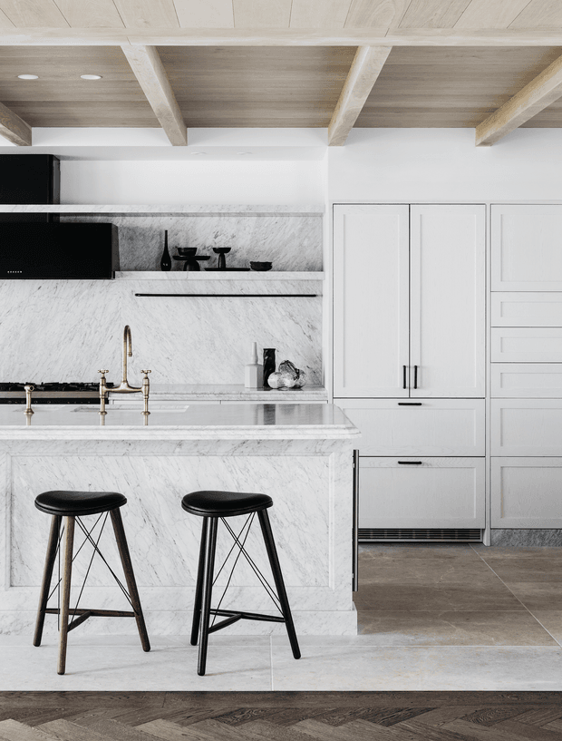 A refined contemporary kitchen featuring extensive white-veined marble cladding on the island and splashback, white shaker-style cabinetry, dark appliances, brass-toned tapware, and black counter stools. The room has a pale timber ceiling with exposed beams, recessed lighting, large-format grey-brown floor tiles, and a darker timber herringbone floor at the foreground transition.