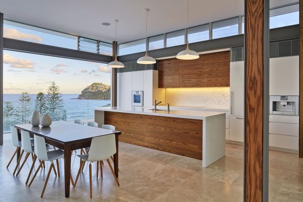 A bright, open-plan kitchen and dining area with expansive floor-to-ceiling glazing overlooking the ocean and a rocky coastal headland. The kitchen features a large white island with a timber-clad base, warm timber overhead cabinetry, pale stone surfaces, integrated appliances, and three white pendant lights. A dark timber dining table with white chairs occupies the foreground, while a prominent timber structural post frames the right side.