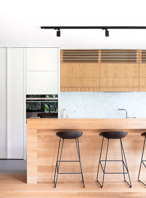 Bright contemporary kitchen featuring a large light timber island with integrated seating, white full-height cabinetry, timber overhead cabinetry with slatted ventilation detailing, and a pale stone or textured splashback. Black track lighting and black bar stools provide contrast. A glimpse of greenery is visible through the rear glazing.