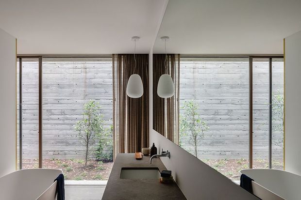 A compact contemporary kitchen viewed from the dining side, centred on a dark stone island or benchtop with an integrated sink and minimalist pendant lights. Full-height glazing frames a planted courtyard enclosed by a horizontal timber privacy fence. A sculptural angled partition and vertical timber detailing add architectural interest, while pale walls and restrained furnishings create a calm, refined atmosphere.