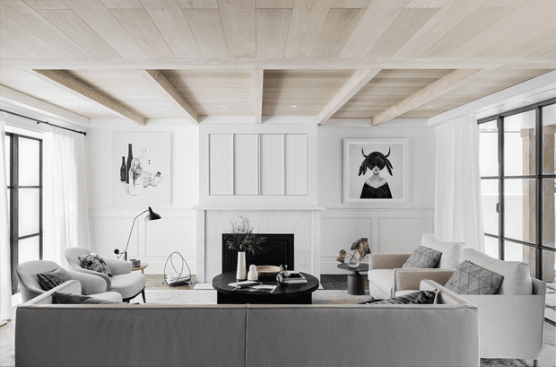 Bright, refined living room with a large sectional sofa facing a black circular coffee table and a white fireplace. The room features extensive white-painted wall panelling, tall black-framed windows with sheer curtains, framed artwork, and a striking light timber coffered ceiling with exposed beams. Neutral furnishings and monochromatic decor create a calm, contemporary Hamptons-inspired interior.