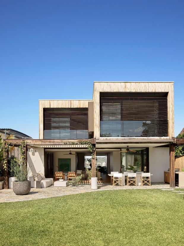 A contemporary two-storey residential house viewed from the landscaped rear garden. The facade combines pale rendered walls with box-like upper volumes clad in vertically textured timber, large glazed openings, and dark horizontal external sun louvres. A covered ground-floor outdoor living and dining area opens onto a paved terrace and lawn, with climbing greenery, planters, and outdoor furniture. The scene is bright and relaxed under a clear blue sky.