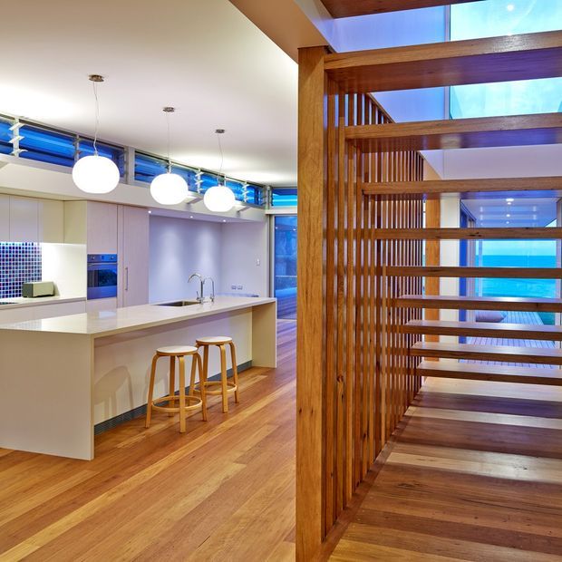 A bright contemporary open-plan kitchen and stairway interior. The kitchen features a large pale island with an integrated sink, light cabinetry, a tiled mosaic splashback, pendant globe lights and timber flooring. In the foreground, a substantial timber stair and slatted timber balustrade create a strong architectural screen, with blue-tinted glazing and a skylit upper level visible beyond.