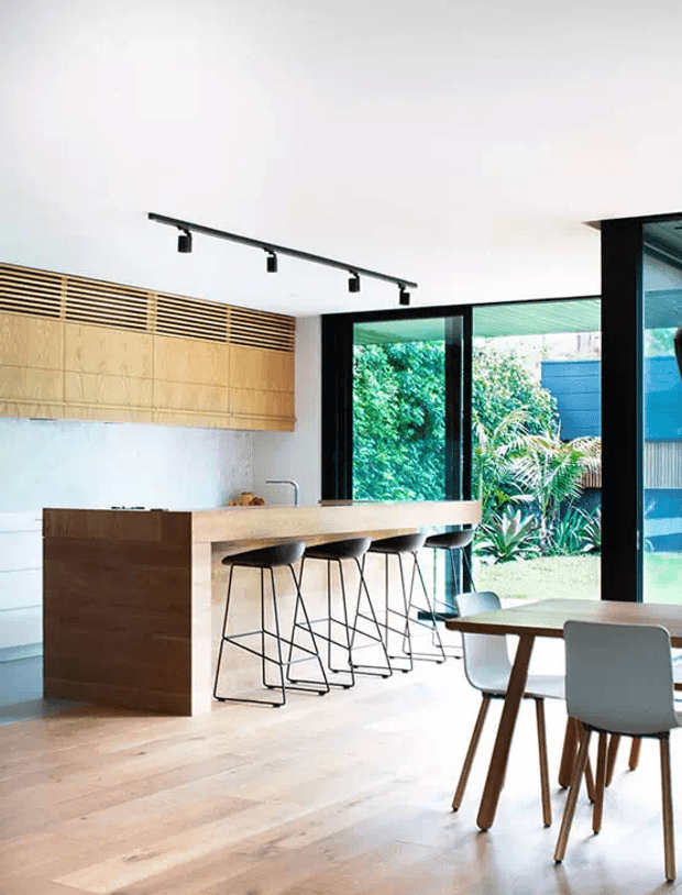 Bright contemporary open-plan kitchen and dining area with pale timber flooring, a large timber island with integrated seating, streamlined timber wall cabinetry, black-framed floor-to-ceiling glazing, and a compact dining table. The space opens onto a lush garden and uses minimalist black track lighting and restrained natural materials.