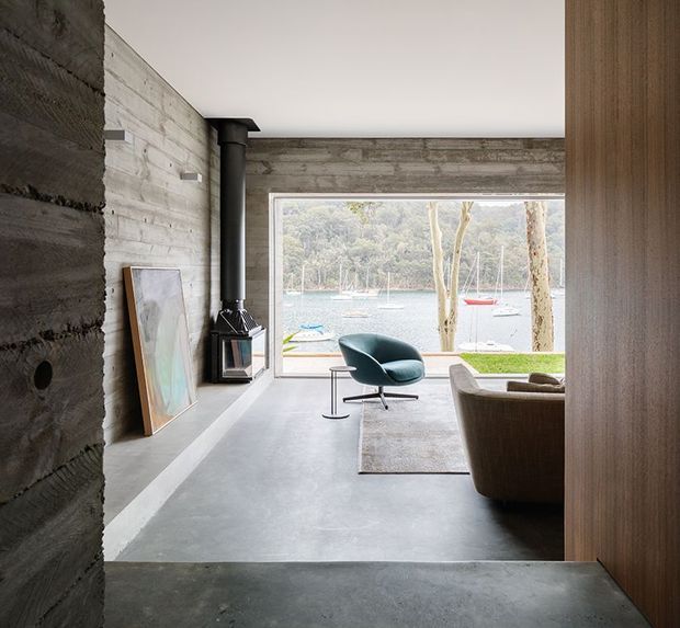 A minimalist living room with raw concrete walls and flooring, a black freestanding wood-burning stove, timber wall panelling, and a large picture window overlooking a waterside landscape. A sculptural teal lounge chair, neutral rug, and brown sofa create a restrained, contemporary retreat-like interior.