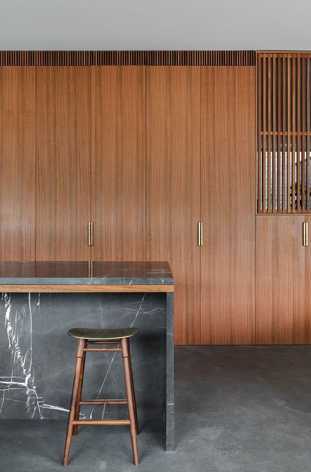 A refined kitchen interior dominated by full-height warm timber cabinetry with slim vertical grain and brass-toned pulls. A dark veined stone island with a timber edge occupies the foreground, paired with a single wooden bar stool. The room has a dark grey polished concrete floor and a vertical timber slatted screen/window detail at right, creating a warm, contemporary and sophisticated atmosphere.
