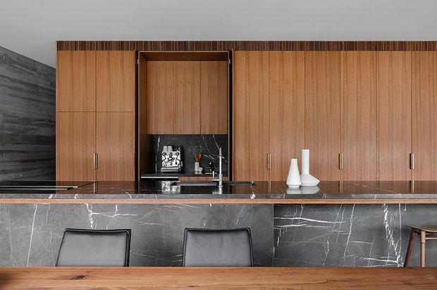 A sophisticated contemporary kitchen and dining area featuring extensive vertical timber cabinetry, a dark marble island with integrated sink and cooktop, and black leather bar seating. The cabinetry includes a recessed dark stone-backed display niche, while a timber dining table occupies the foreground. Dark textured wall finishes and contrasting white decorative vessels add depth to the restrained palette.