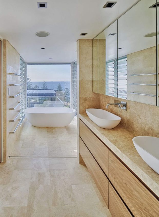 Bright contemporary bathroom with a freestanding white bathtub positioned beside full-height louvred glazing overlooking a coastal view. The room features large beige natural-stone wall and floor surfaces, a matching stone vanity benchtop, extensive mirrored cabinetry, pale timber vanity joinery, twin vessel basins, recessed ceiling lighting, and open shelving.