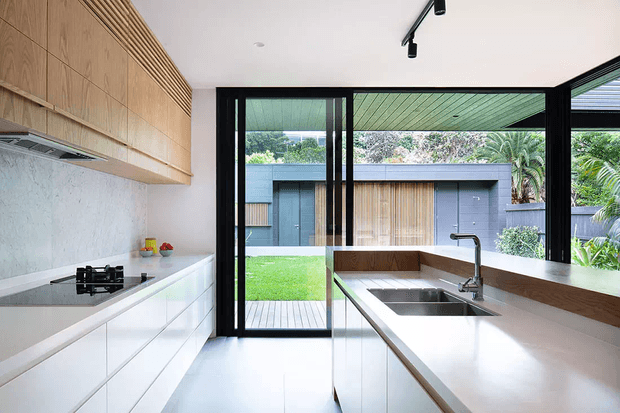 Bright contemporary kitchen with white lower cabinetry, warm timber overhead cabinetry and a timber-lined backsplash, a large white island with integrated stainless-steel sink, black-framed sliding glass doors, and a view to a landscaped courtyard and adjoining modern pavilion.