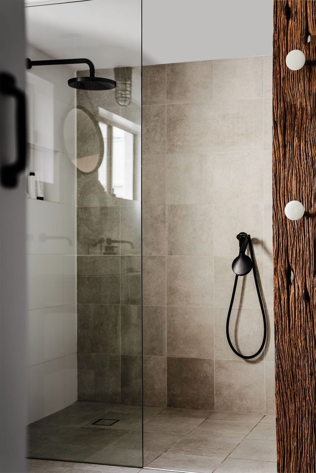 A close-up view of a contemporary walk-in shower enclosed by frameless glass. The shower has large beige-grey stone-look wall and floor tiles, matte black ceiling-mounted and handheld fixtures, a linear drain, and a warm textured timber feature wall with round white controls or fittings at the right edge. The glass reflects the shower interior and a circular mirror or feature beyond.
