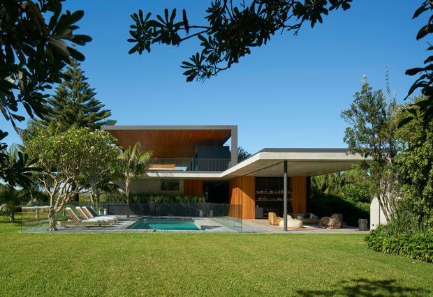 A contemporary two-storey residence viewed from a landscaped lawn, with strong horizontal roof planes, extensive glazing, warm timber soffits and cladding, and a covered outdoor entertaining area beside a swimming pool. Tropical and subtropical planting frames the house, while a broad flat roof shelters an open lounge or dining area.