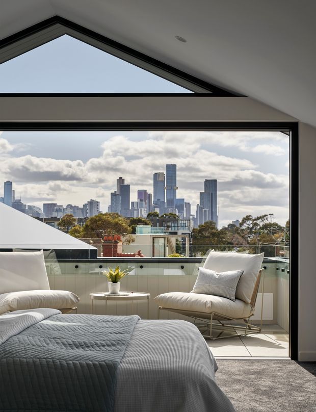 Bright contemporary bedroom with a large corner window and angled clerestory glazing overlooking a dense city skyline. The room features a soft upholstered bed, light grey textured bedding, a matching lounge chair, a small round side table, carpeted flooring, and a glazed balcony beyond the windows.