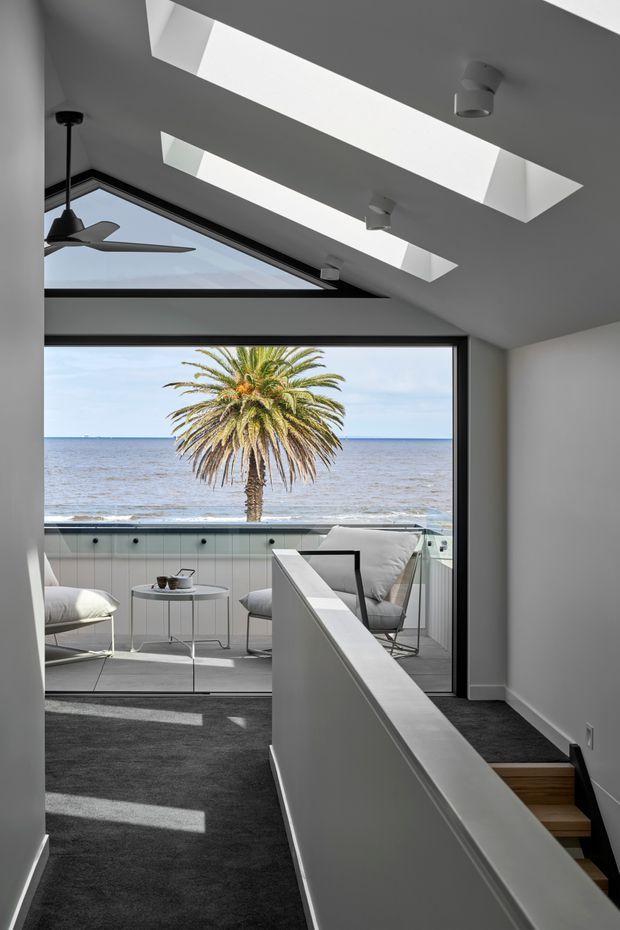 A bright, minimalist interior landing or hallway overlooking a seaside balcony. The space features a dark carpeted floor, white walls and ceiling, recessed skylights, a ceiling fan, black-framed floor-to-ceiling glazing, and a white balustrade beside a short timber stair. Through the glazing, a furnished balcony with pale built-in paneling and outdoor seating faces the ocean and a palm tree.