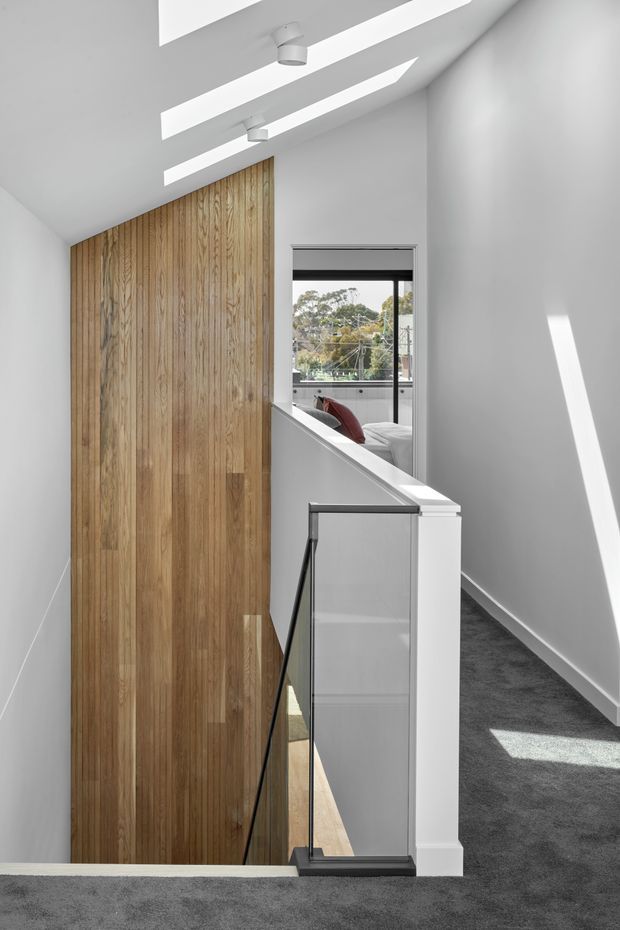 Bright contemporary upper-level hallway and stair landing with a dramatic sloped ceiling, linear skylights, white walls, dark grey carpet, and a tall vertical timber feature wall beside the stair void. A minimalist glass-and-dark-metal balustrade overlooks the stairs, with a bedroom visible through the opening at the far end.